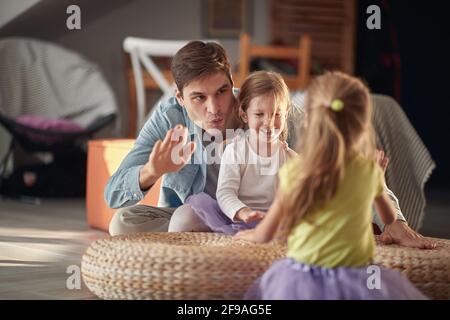 Ein junger Vater in fröhlicher Atmosphäre zu Hause lehrt seine Töchter, wie man auf die Möbel trommeln. Familie, Zuhause, Spielen, Zweisamkeit Stockfoto