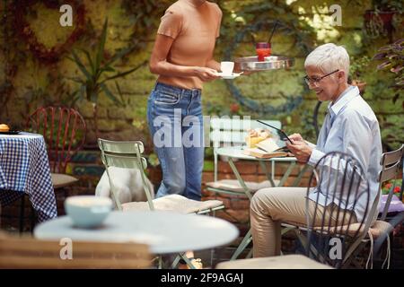 Eine ältere Frau, die in entspannter Atmosphäre an der Bar ein Tablet benutzt, während sie einen Kaffee getrunken hat. Freizeit, Bar, Freundschaft, Outdoor Stockfoto