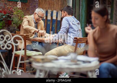 Ein älterer Mann, der sich mit seinem jungen Freund vergnügt, während er in einer fröhlichen Atmosphäre in der Bar Schach spielt. Freizeit, Bar, Freundschaft, Outdoor Stockfoto