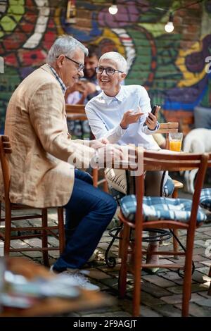Ein älteres Paar schaut sich Smartphone-Inhalte an, während es in entspannter Atmosphäre in der Bar einen Drink zu sich nehmen kann. Freizeit, Bar, Freundschaft, Outdoor Stockfoto