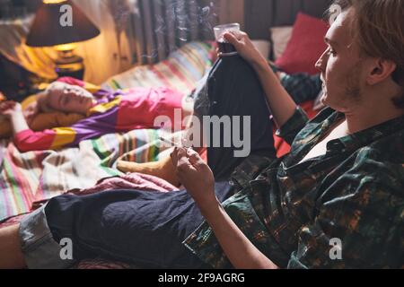 Junger Mann, der eine Zigarette raucht und Wein trinkt Das Bett mit seiner Freundin, die sich in der Nähe von ihm entspannt hat Zu Hause Stockfoto