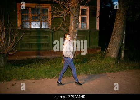 Ein schönes junges Mädchen in Jeans und einer beigen Jacke posiert auf der Veranda eines typischen russischen Supermarkts. Stockfoto