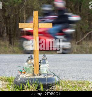 Ein Kreuz am Straßenrand mit Kerzen zum Gedenken an den tragischen Tod, auf einer Fahrt im Hintergrund verschwommenes Motorrad Stockfoto