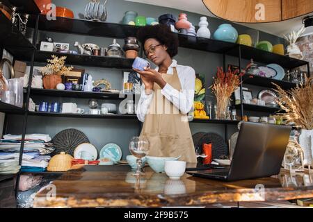 Erfahrene afrikanische Verkäuferin, die in einem modernen Laden mit verschiedenen Dekorationen Inventar anführt. Junge Dame mit Brille und Schürze, die während des Arbeitsprozesses einen kabellosen Laptop benutzt. Stockfoto