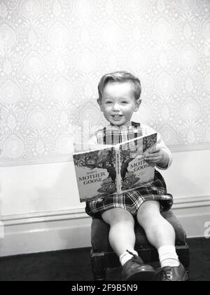 1960er Jahre, historisch, in einem Raum, ein glücklicher kleiner Junge in einer Tartan gemusterten Latzhose, sitzend auf einem Hocker mit dem Buch "Baby's Mother Goose", England, Großbritannien. Mother Goose ist die imaginäre Autorin einer Sammlung französischer Märchen und später populärer englischer Kinderreime. Stockfoto