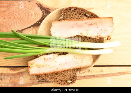 Zwei Scheiben köstlicher, kalorienreicher, leckerer, pikanter Speck mit Brot und frischen Zwiebeln auf einer Keramikuntertasse, die auf einem Holztisch steht. Stockfoto