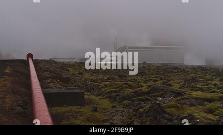 Blick auf das Kraftwerk Svartsengi in der Nähe von Grindavik, Halbinsel Reykjanes, Island mit moosbedecktem Lavafeld vor der Tür und rot gefärbter Pipeline. Stockfoto