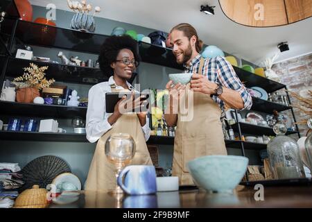 Zwei multirassische Partner, die digitale Tablets verwenden, während sie Waren im Einrichtungsgeschäft empfangen. Afrikanische Frau und kaukasischer Mann tragen beige Schürzen, arbeiten zusammen. Stockfoto