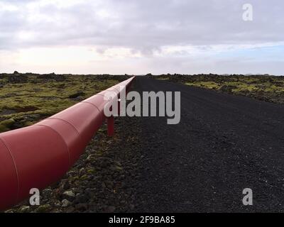 Abnehmende Perspektive der rot gefärbten Metallpipeline neben einer Schotterstraße aus schwarzen Steinen, umgeben von moosbedeckten Lavafeldern bei Svartsengi. Stockfoto