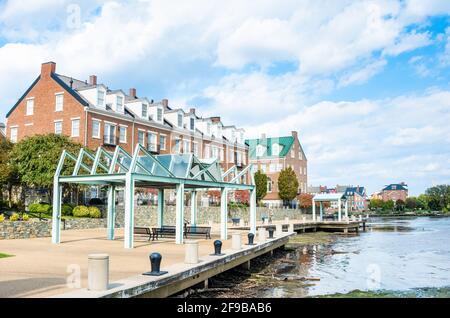 Metallbänke und stilvolle Pavillons an einem sonnigen Herbstmorgen entlang eines verlassenen Spazierweges am Flussufer. Moderne Backsteinhäuser sind im Hintergrund zu sehen. Stockfoto