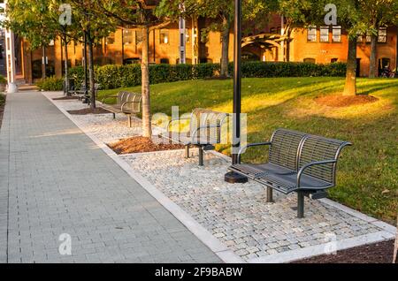 Leere moderne Stahlbänke entlang eines gepflasterten Fußweges in einem Öffentlicher Park, der bei Sonnenuntergang im Herbst von Backsteingebäuden umgeben ist Stockfoto