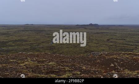 Luftpanorama über den südlichen Teil der Halbinsel Reykjanes, Island mit spärlicher Landschaft aus moosbedeckten Lavafeldern mit Straße und Pipeline. Stockfoto