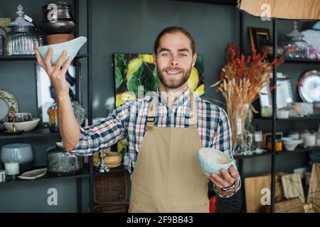 Professioneller männlicher Verkäufer mit exklusiver Careamic Tasse und Vase in den Händen. Ein hübscher bärtiger Mann mit beigefarbener Schürze empfängt Waren im modernen Einrichtungsgeschäft. Stockfoto