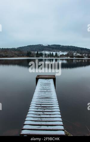 Leerer hölzerner Pier am See, der mit Neuschnee bedeckt ist. Winterteich mit kleinem Steg und Schnee. Nebliger See mit Hügeln. Weiße Winterlandschaft.Bewölkt, kalt Stockfoto