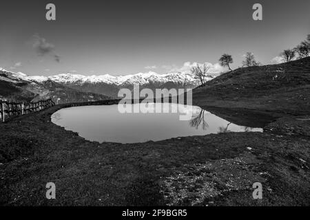 Graustufenaufnahme eines Teiches auf einem Bergfeld mit verschneiten Bergen Im Hintergrund Stockfoto