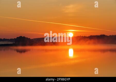 Der wunderschöne Sonnenuntergang spiegelt sich auf dem See wider. Himmel mit orangefarbenen und gelben Wolken. Majestätischer Frühlingsmorgen. Nebelwald. Stockfoto