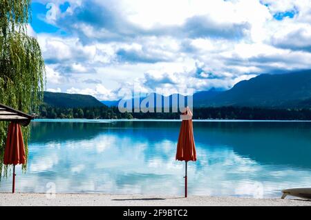 Leere Strandbar auf einem Bergsee, Blick über das Wasser mit Wolkenreflexen, zwei malerische Sonnenschirme im Vordergrund
