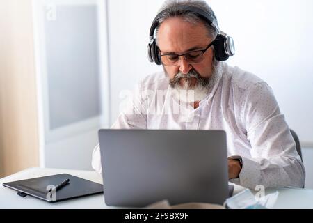 Ein älterer Mann mit Kopfhörer, der eine Videokonferenz mit einem Laptop veranstaltet Zu Hause Stockfoto