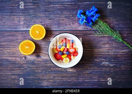 Gesunde Frühstückschale mit Cerealien und frischen Früchten (Erdbeere, Banane, Heidelbeere, Mango) und Orangenfrüchten auf einem hölzernen Frühstückstisch. Stockfoto