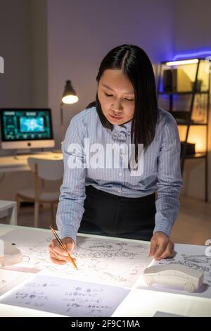 Konzentrierter junger asiatischer Automobildesigner, der im Dunkeln am Tisch steht Büro und Arbeiten mit Layouts von Fahrzeugkomponenten Stockfoto