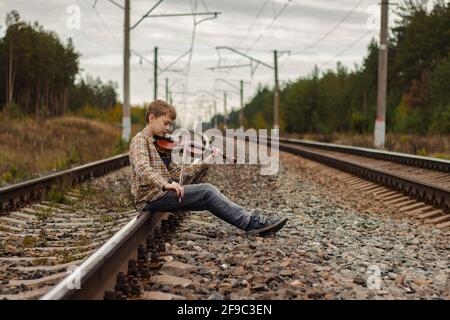 Ein schöner blonder Junge sitzt auf den Schienen der Eisenbahn und spielt Geige. Stockfoto