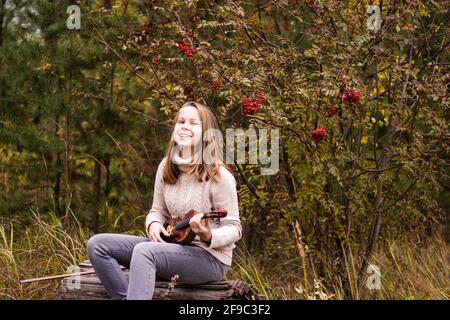 Ein wunderschönes Mädchen im Teenageralter mit langen blonden Haaren sitzt auf einem Baumstamm im Park und hält eine Geige in den Händen, sieht happ aus Stockfoto