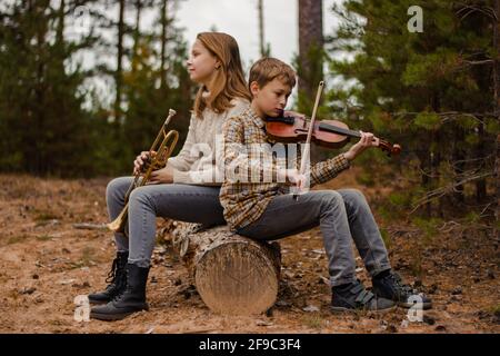 Junge und Mädchen, Bruder und Schwester, Teenager spielen Trompete und Geige, die im Wald auf einem Holzstamm sitzen. Stockfoto