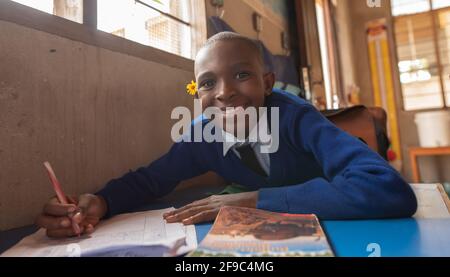 Dodoma, Tansania. 08-18-2019. Porträt eines schönen schwarzen Mädchens, das lächelt und eine Blume auf ihrem Hören hat und auf die Kamera schaut, während sie auf einer Bank sitzt Stockfoto