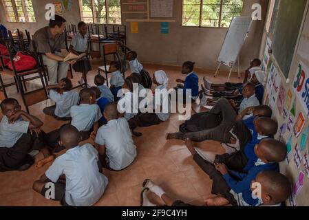 Dodoma, Tansania. 08-18-2019. Weitwinkelaufnahme mit schwarzen Kindern, die in ihrem Klassenzimmer auf dem Boden sitzen und dem Vortrag ihres Lehrers in einem ländlichen sc lauschen Stockfoto