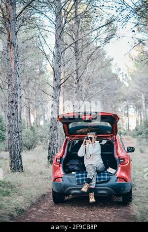 Junge Erwachsene kaukasische Frau, die im Herbst in der Nähe eines Autos fotografiert, das auf einem Forstweg geparkt ist. Reisekonzept. Stockfoto