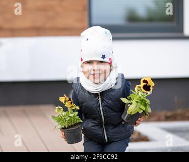 Foto eines kleinen Mädchens, das Stiefmütterchen in Töpfen in den Händen hält, die Assistentin der Mutter. Stockfoto