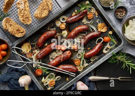 Blick von oben auf köstliche gegrillte Würstchen, serviert mit Zwiebeln, Tomaten, Knoblauch, Brot und Kräutern Stockfoto