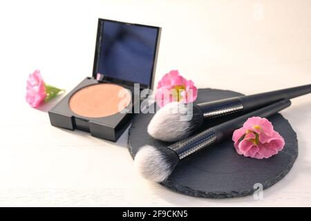 Kosmetische Pinsel auf einem Schiefer, Make-up Gesicht Pudercase und rosa Blumen auf einem weißen Holzhintergrund, Beauty-Konzept, Kopierraum, ausgewählten Fokus, sehr Stockfoto