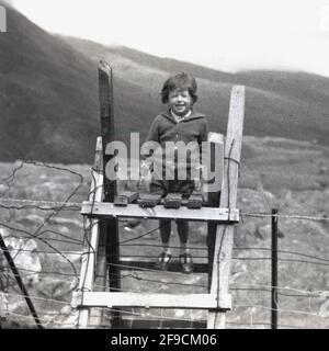 1960s, historisch, draußen in den Yorkshire Dales, ein junges Mädchen, das auf den Stufen eines Holzstelns auf einem Stacheldrahtzaun steht. Ein Stil ist eine Reihe von Schritten, die es einem ermöglicht, über einen Zaun oder eine Barriere zu gelangen, d. h., von einem Feld zum nächsten zu übertragen, aber verhindert, dass alle Nutztiere oder Tiere entkommen, England, Großbritannien. Stockfoto