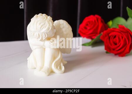 Eine kleine Skulptur eines kleinen Engels mit Flügeln und einer roten Rose auf schwarzem Hintergrund. Das Konzept von Beerdigungen, Beileid und Trauer. Stockfoto