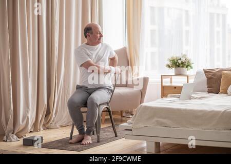 Sportlicher älterer Mann, der mit einem Stuhl Yoga praktiziert, Parivrtta Utkatasana macht, im Wohnzimmer zu Hause trainiert Stockfoto