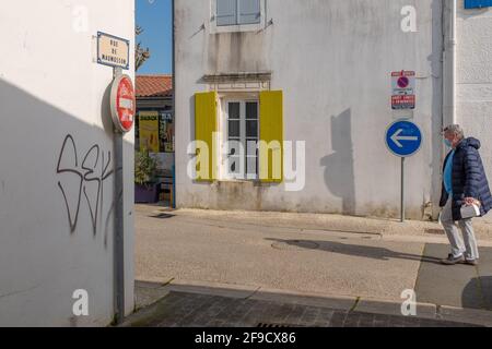 Saint-Trojan-les-Bains, Frankreich - 26. Februar 2020: Eine Person, die eine Maske trägt und einen langen Schatten wirft, geht auf der Straße der Altstadt in der Sonne spazieren Stockfoto