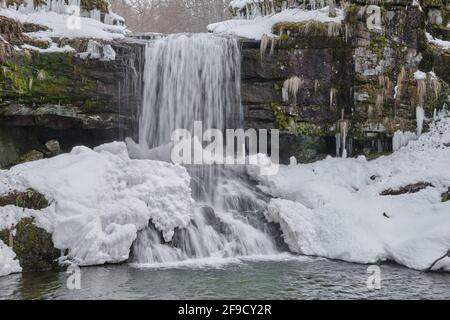 Schneebedeckter, landschaftlich reizvoller Wasserfall, der im Winter die nassen, strukturierten Felsen einer Klippe hinunterstürzt Stockfoto