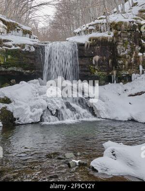 Schneebedeckter, landschaftlich reizvoller Wasserfall, der im Winter die nassen, strukturierten Felsen einer Klippe hinunterstürzt Stockfoto