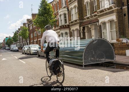 Radfahrer (Bewegung verschwommen), die an einem abschließbaren Fahrradraum auf der Straße vorbeifahren. Stockfoto