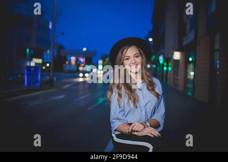 Fröhliche schöne bosnisch kaukasische Frau in Hut posiert auf der Straße mit Stadtlichtern Stockfoto