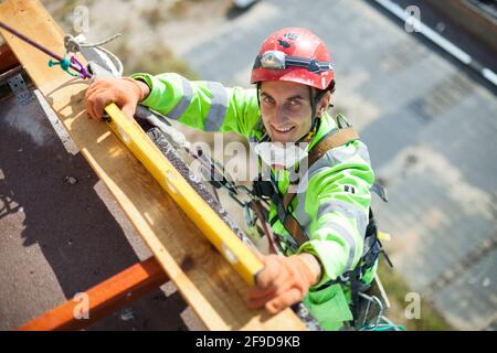 Industriekletterer misst während der Bauarbeiten mit einem Ebenen Rohr, blickt auf die Kamera und lächelt Stockfoto