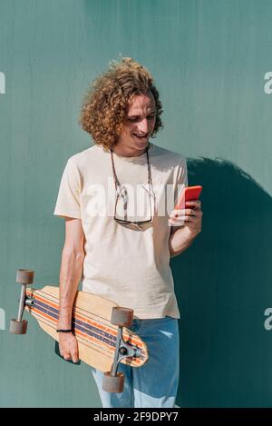 Vertikales Porträt eines langhaarigen jungen Mannes, der sein Telefon überprüft. Er trägt lässige Kleidung und hält ein Skateboard Stockfoto