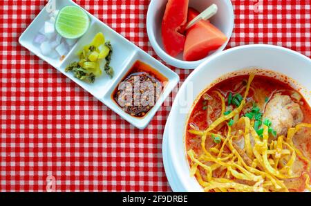 Nördliche Thai-Nudelsuppe mit Huhn und Beilage in weißer Schüssel auf rot karierter Tischdecke. Draufsicht auf Khao Soi und reife Papaya mit Holz Stockfoto