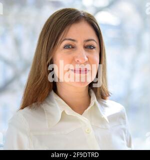 Nachdenkliche Frau mittleren Alters, die leise auf die Kamera schaut Lächeln in der Nähe Kopf und Schultern gegen Ein großes Fenster Stockfoto