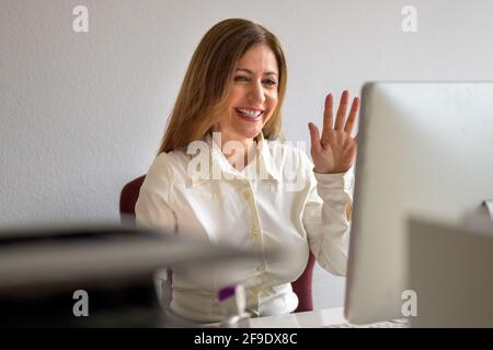 Geschäftsfrau, die bei einem Videokonferenzanruf oder einer Remote-Verbindung online interagiert Arbeitstreffen winken oder heben ihre Hand, um zu wählen und Lachend vor ihrem d Stockfoto