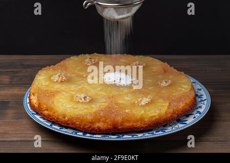 Eine Ananas- und gehackte Mandelkuchen, umgedreht genannt, wird mit Puderzucker bestäubt, der aus einem Sieb fällt Stockfoto