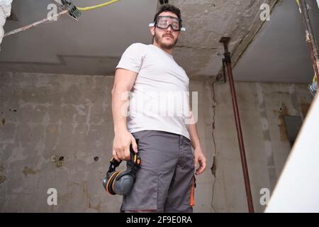 Junger Baumeister mit Schutzbrille auf einer Baustelle. Gehörschutz hängt an der Hose. Stockfoto