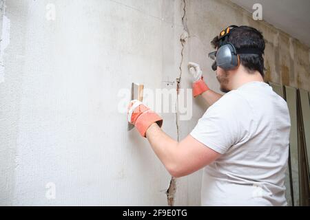 Bauarbeiter repariert einen Riss und verputzte Zement an der Wand. Der Baumeister trägt Schutzhandschuhe, eine Brille und einen Ohrenschützer. Stockfoto