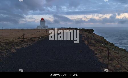 Schwarzer Wanderweg auf der Spitze der Halbinsel Dyrhólaey bei Vík í Mýrdal an der Südküste Islands am Abend mit Leuchtturm und bewölktem Himmel. Stockfoto